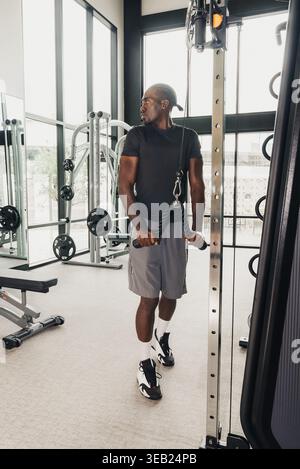 Black Man Coach Geht Durch Das Fitnessstudio. Legere Stride Mit Kabellosen Kopfhörern, Handtuch Über Schulter, Ausgekleideter Flur, Tageslicht, Aktiv Stockfoto