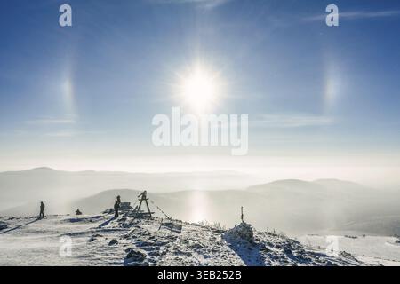 Sonnenhunde über verschneiten Bergketten mit Menschen, die die Winterlandschaft bewundern. Reiseerlebnis bei kaltem Wetter für Erkundungskonzept Stockfoto