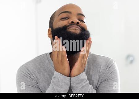 Afrikanischer Mann, der Bart mit beiden Händen schröpft, im Badezimmer schaut und ein hellgraues Langarmhemd trägt Stockfoto