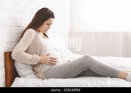 Schwangere Frau, die sich beim Sitzen auf einem Bett in einem hellen Zimmer unwohl fühlt Stockfoto