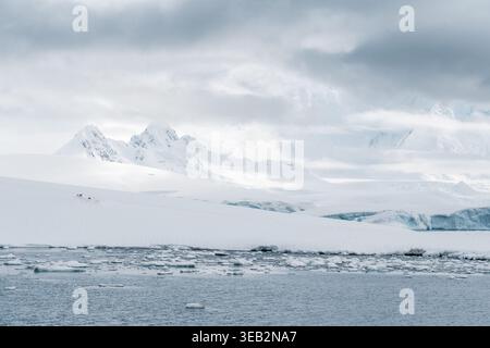 Blick auf schneebedeckte Berggipfel und schwimmendes Eis im kalten Wasser unter einem bewölkten Himmel in der Antarktis. Stockfoto