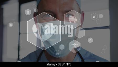 Stehender maskierter Arzt in Operationskappe und Peelings mit Stethoskop in der Klinik, mit Hex-Ikonen Stockfoto