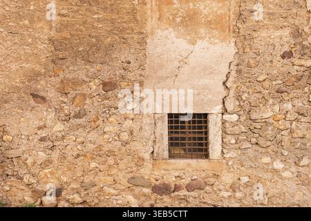 Blick auf eine alte strukturierte Steinmauer mit einem kleinen quadratischen Fenster mit einem rostigen Eisengitter und verwittertem Putz Italien. Stockfoto