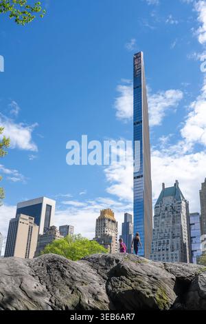 Blick auf den schlanken Steinway Tower, der sich über dem üppigen Grün und den grauen Felsen des Central Park erhebt, mit Kindern, die in New York, New York, Vereinigte Staaten spielen Stockfoto