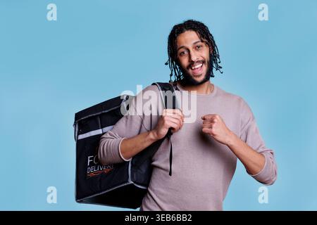 Fröhlicher Kurier mit Essenszustellung, der mit einem Thermorucksack und einem lächelnden Porträt steht. Junger sorgloser Zustellant, der Essen zum Mitnehmen in der Tasche liefert und in die Kamera schaut Stockfoto