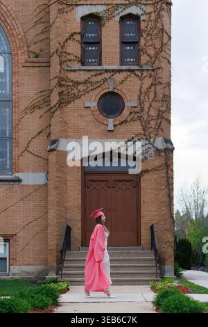 Nach der Abschlussfeier spaziert ein Student in Mütze und Kleid an einem historischen Gebäude vorbei. Der Schüler konzentriert sich auf den Eingang und feiert Stockfoto
