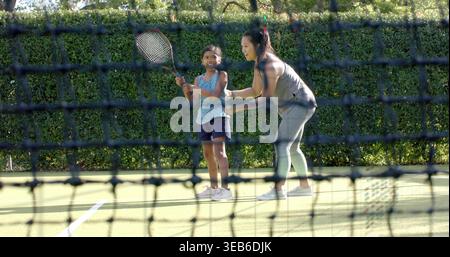 Frau, die ein Mädchen führt, das Tennisschläger auf dem Tennisplatz hält, in Turnschuhen, mit Netz und Basketballkorb Stockfoto