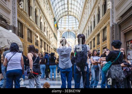 Eine Gruppe von Touristen von hinten, mit einem Mann in einem karierten Hemd, die ein Foto von der monumentalen Kuppel in der Galleria Umberto I. Neapel, Italien - 17. April 2026: Mehrere Touristen stehen im Mittelschiff der Galleria Umberto I. Ein Mann in einem karierten Hemd hält ein Smartphone hoch, um ein Foto von der Glaskuppel zu machen. Die Szene wird durch natürliches Licht beleuchtet, das durch die Eisenstruktur gefiltert wird. Neapel NA Italien Copyright: XGennaroxLeonardix Stockfoto