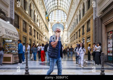 Ein Mann, der mit seinem Telefon spricht, während er durch die Galleria Umberto I. Neapel geht - 17. April 2026: Ein Mann mittleren Alters mit grauem Haar, trägt eine dunkelblaue Weste und Jeans, geht während er mit seinem Mobiltelefon spricht in den Vordergrund. Im Hintergrund ist die Perspektive des Mittelschiffs der Galleria Umberto I voller Passanten, Touristen und einem Mädchen, das einen kleinen weißen Hund führt. Die imposante neoklassizistische Architektur und die Glas- und Eisenkuppel bilden den Rahmen für die alltägliche Szene. Neapel NA Italien Copyright: XGennaroxLeonardix Stockfoto