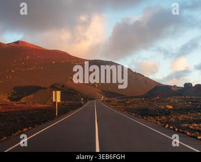 Blick auf eine leere Asphaltstraße, die sich durch die vulkanische Landschaft des Timanfaya Nationalparks mit rötlichen Bergen unter dem weichen Sonnenuntergang Lanzaro schlängelt Stockfoto