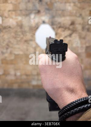 Aus erster Sicht eines Mannes, der eine schwarze halbautomatische Pistole mit einer Hand auf ein weißes Ziel gegen eine Ziegelmauer zielt. Stockfoto