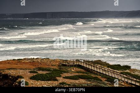 Portugals atlantikküste erlebt starke Winde und mächtige Meereswellen, die unter einem bewölkten Himmel mit einem Holzbrett gegen die Küste brechen Stockfoto