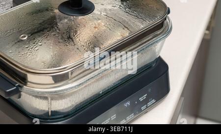 Glasbehälter mit schwarzem Deckel auf dem Induktionskochfeld, der während des Kochens Kondenswasser im Inneren zeigt. Hausküche kochen, Speisen dampfen im Behälter Stockfoto