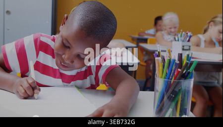 Schreibendes Kind in rot gestreiftem T-Stück, das sich über Papier am Klassenpult gelehnt hat, mit durchsichtigem Bleistiftbehälter Stockfoto