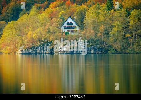 Blick auf ein weißes Haus eingebettet zwischen lebhaften Herbstbäumen am felsigen Ufer des Bohinj-Sees mit ruhigen Wasserspiegeln Bohinjsko jezero, Radovljica, S Stockfoto