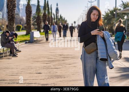 Batumi, georgia 21. april 2026: Junge Frau auf der batumi Promenade konzentrierte sich auf Smartphone, ging auf palmengesäumten Stadtpfaden, schrieb SMS und stöberte im Freien Stockfoto