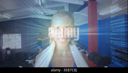 Lächelnde Frau in athletischer Spitze, die im Fitnessstudio unter Balken steht, weißes Handtuch, holographischer Code-Überzug Stockfoto