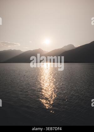 Blick auf eine Person, die auf einem Paddleboard inmitten eines ruhigen Sees steht und die Sonne hinter Berggipfeln in Kanada untergeht. Stockfoto