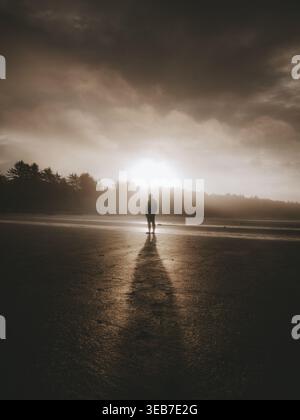 Blick auf eine Person, die an einem Sandstrand unter einem dramatischen bewölkten Himmel bei Sonnenuntergang in Kanada steht. Stockfoto
