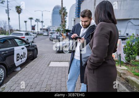 Batumi, georgia 21. april 2026: Mann bestellt Bolt Ride in batumi mit Smartphone-App, während die Frau auf dem belebten städtischen Bürgersteig wartet, tagsüber Stadttransporte Stockfoto