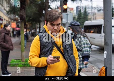 Batumi, georgia 21. april 2026: Junger Mann mit gelbem Hoodie und schwarzer Weste, überprüft sein Smartphone und hört Musik in der Stadt batumi Stockfoto