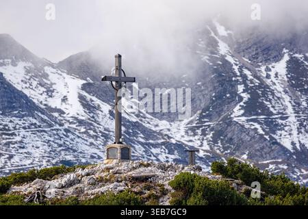 Blick auf das hölzerne Gipfelkreuz mit einer Glocke im Skizentrum Vogel auf einem felsigen Gipfel mit schneebedeckten Bergen und nebeligen Wolken in Ukanc, Radovljica, Slov Stockfoto