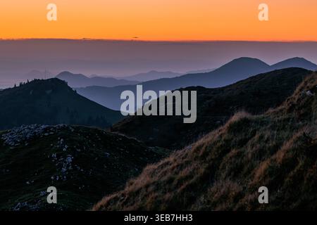 Blick auf mehrschichtige Bergsilhouetten unter einem leuchtend orangefarbenen Sonnenuntergangshimmel mit nebeligen Tälern in Sorica, Skofja Loka, Slowenien. Stockfoto