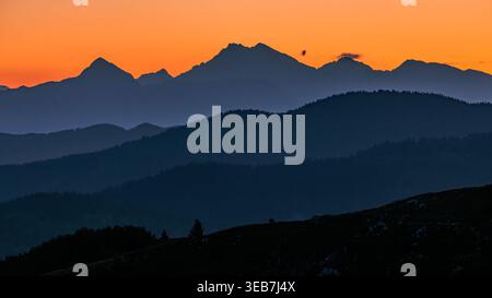 Blick auf mehrschichtige Bergsilhouetten unter einem leuchtend orangefarbenen Himmel bei Sonnenuntergang Sorica, Skofja Loka, Slowenien. Stockfoto