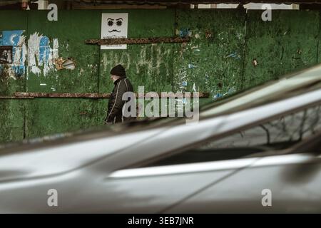 Batumi, georgia 21. april 2026: Ein älterer Mann in warmen Kleidern geht auf einer Stadtstraße in batumi, georgia, mit einem leuchtend grünen, strukturierten Wandbehi Stockfoto