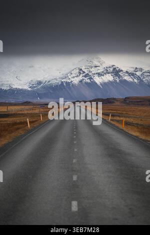 Blick auf eine lange Asphaltstraße mit einem Auto in der Ferne, das sich in Richtung eines majestätischen, schneebedeckten Berges unter einem dunklen, stimmungsvollen Himmel in Island erstreckt. Stockfoto