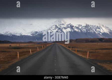 Blick auf eine gerade Asphaltstraße, die sich in Richtung massiver schneebedeckter Berge unter einer schweren dunklen Wolkenschicht in Island erstreckt. Stockfoto
