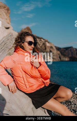 Frau Pufferjacke Strand junge Frau mit einem leuchtend orangen Mantel und Sonnenbrille, die an einem felsigen Ufer am Meer sitzt Stockfoto