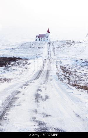 Blick auf die historische Snaefellsnes-Kirche mit ihrem roten Dach am Ende einer langen schneebedeckten Straße, die sich durch eine weiße Winterlandschaft schlängelt Stockfoto