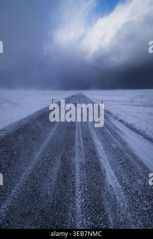 Blick auf eine gerade, mit Schnee und Eis bedeckte Asphaltstraße, die sich durch eine weite Winterlandschaft unter einem dramatischen bewölkten Himmel in Snaefellsnes, Eyja- Stockfoto