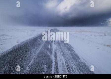 Blick auf eine schneebedeckte Asphaltstraße, die in Richtung dunkler Sturmwolken über die weiße Tundra von Snaefellsnes, Eyja- og Miklaholtshr, Island, führt. Stockfoto