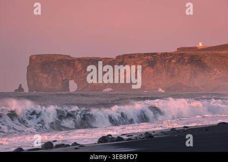 Blick auf den Felsbogen Dyrholaey und den Leuchtturm bei Sonnenuntergang mit stürzenden Wellen am schwarzen Sandstrand in Vik, Myrdalshreppur, Island. Stockfoto