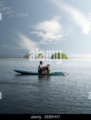 Blick auf eine Person in einem kleinen blauen Boot auf dem Amazonas, die aus einem Container mit grünen Inseln unter einem bewölkten Himmel strömt. Stockfoto