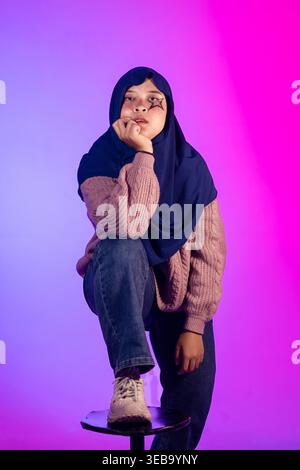 Eine junge Frau im Hijab posiert nachdenklich. Der violette und pinkfarbene Hintergrund sorgt für mehr Lebendigkeit. Sie sitzt auf einem kleinen Hocker mit einem überkreuzten Bein Stockfoto