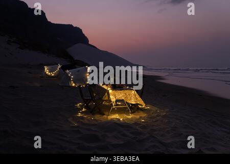 Blick auf einen Strandcamping mit beleuchteten Zelten und einem Esstisch unter einem violetten Abendhimmel in Socotra, Jemen. Stockfoto