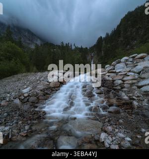 Felsiger Alpenbach, der von einem kleinen Wasserfall unter nebeligen bewaldeten Bergen in Tirol, Österreich, fließt. Stockfoto