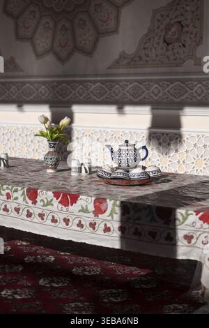 Blick auf ein traditionelles Teeset aus Keramik und eine Vase mit Blumen auf einer gestickten Tischdecke in einem reich verzierten Zimmer in Taschkent, Usbekistan. Stockfoto