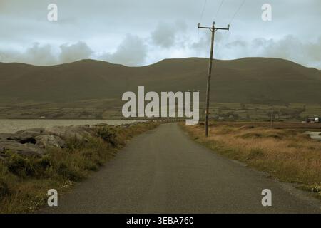 Eine leere malerische Küstenstraße, die in Richtung dunkler Berge unter einem bedeckten Himmel in Irland führt. Stockfoto