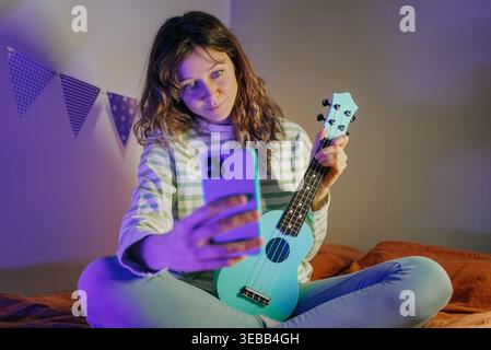 Junge Frau, die Inhalte erstellt und einen Livestream macht, ein Selfie-Video mit Ukulele und Smartphone für soziale Medien aus ihrem Schlafzimmer macht, beleuchtet von sanftem Blau- und violettem Neonlicht Stockfoto