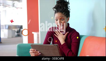 Frau in kastanienbraunem Pullover halten, als Reaktion auf Tablette auf dem türkisfarbenen Sofa im Büro Stockfoto