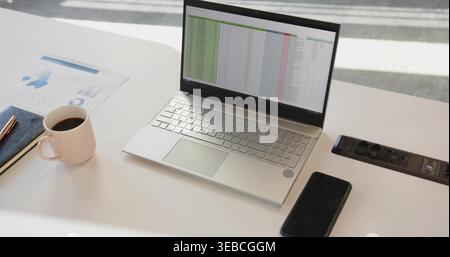 Tabellenkalkulation, offenes silbernes Notebook in der Mitte des weißen Schreibtisches, Kaffeetasse und Smartphone Stockfoto