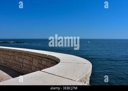 Minimal geschwungene Steinmauer mit Blick auf das tiefblaue Meer und den Horizont unter klarem Himmel. Mediterraner Küstenhintergrund mit großem Kopierraum für Design Stockfoto