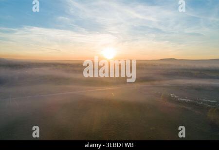 Die helle Sonne aus der Vogelperspektive besticht durch eine nebelige Tallandschaft und wirft bei Sonnenaufgang warmes Licht über grüne Felder und ferne Berge Stockfoto