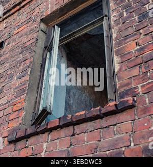 Ein alter Holzfensterrahmen in einer verwitterten roten Ziegelwand des heruntergekommenen Gebäudes zeigt einen dunklen Innenraum Stockfoto