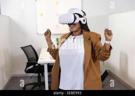 Afroamerikanerin im mittleren Erwachsenenalter mit Kamelblazer und VR-Headset, stehend am Arbeitsplatz Stockfoto