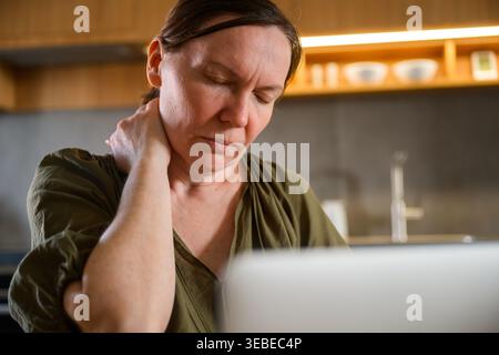 Reife Frau mit geschlossenen Augen, die den Nacken in der Nähe des Laptops reibt, Müdigkeit und Muskelverspannungen durch Computerarbeit. Selektiver Fokus. Stockfoto